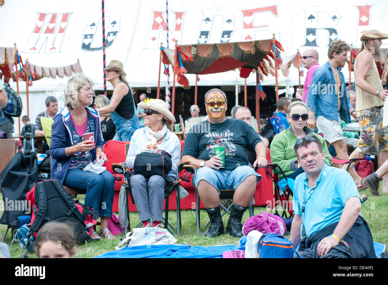 Middle-aged man with his face painted as a tiger and his mates drinking beer sitting at a music festival by flags and tents Stock Photo