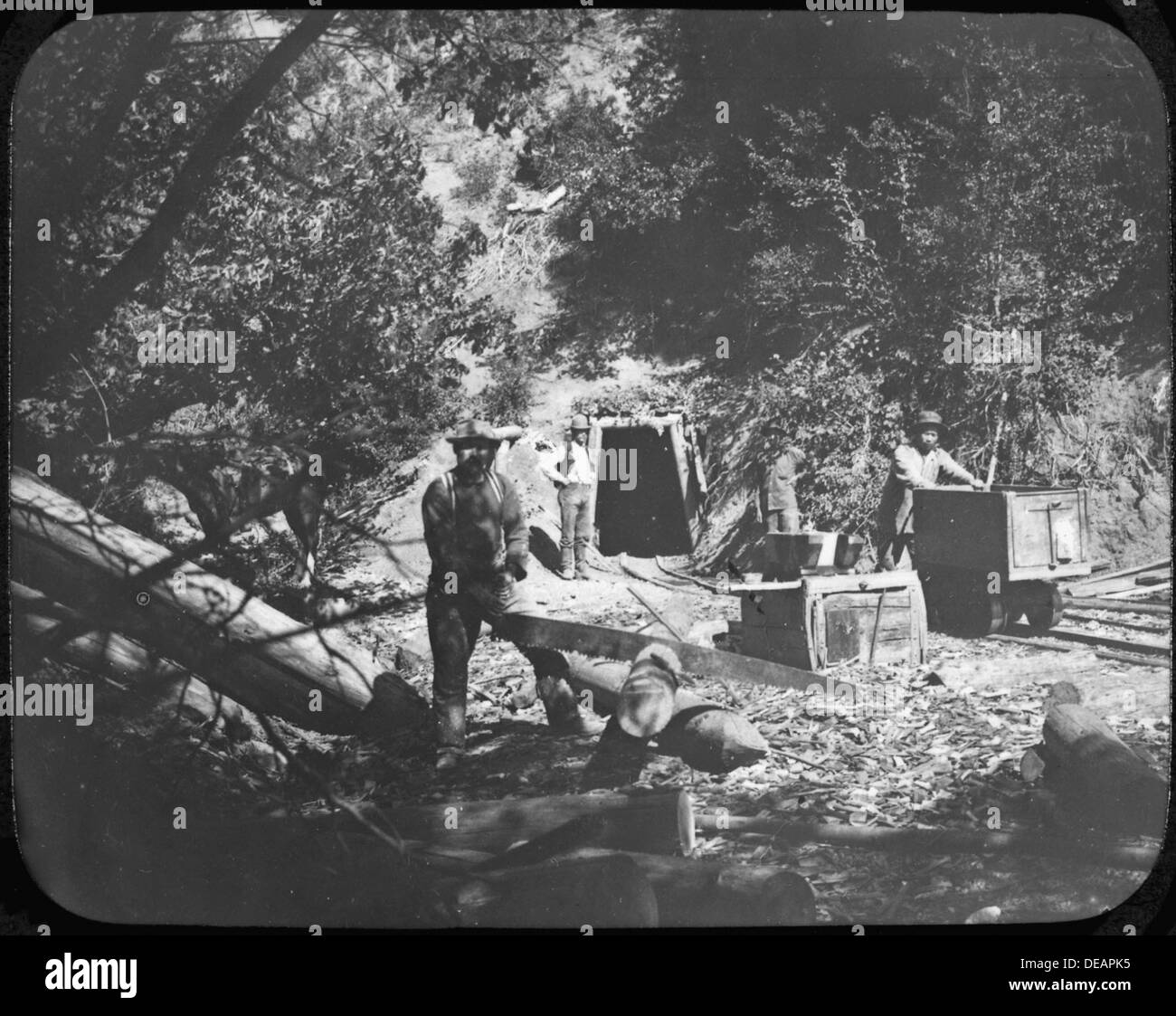 Photograph of four men outside a mine shaft. The foremost man appears to be cutting timbers for bracing the interior 296580 Stock Photo