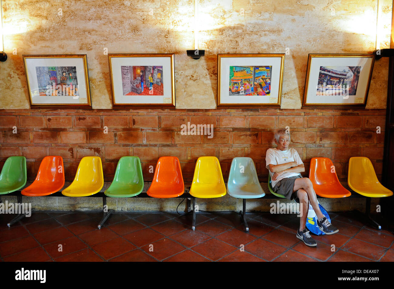 old man wearing shorts and tee shirt sitting on a row of coloured plastic chairs with coloured paintings. Stock Photo
