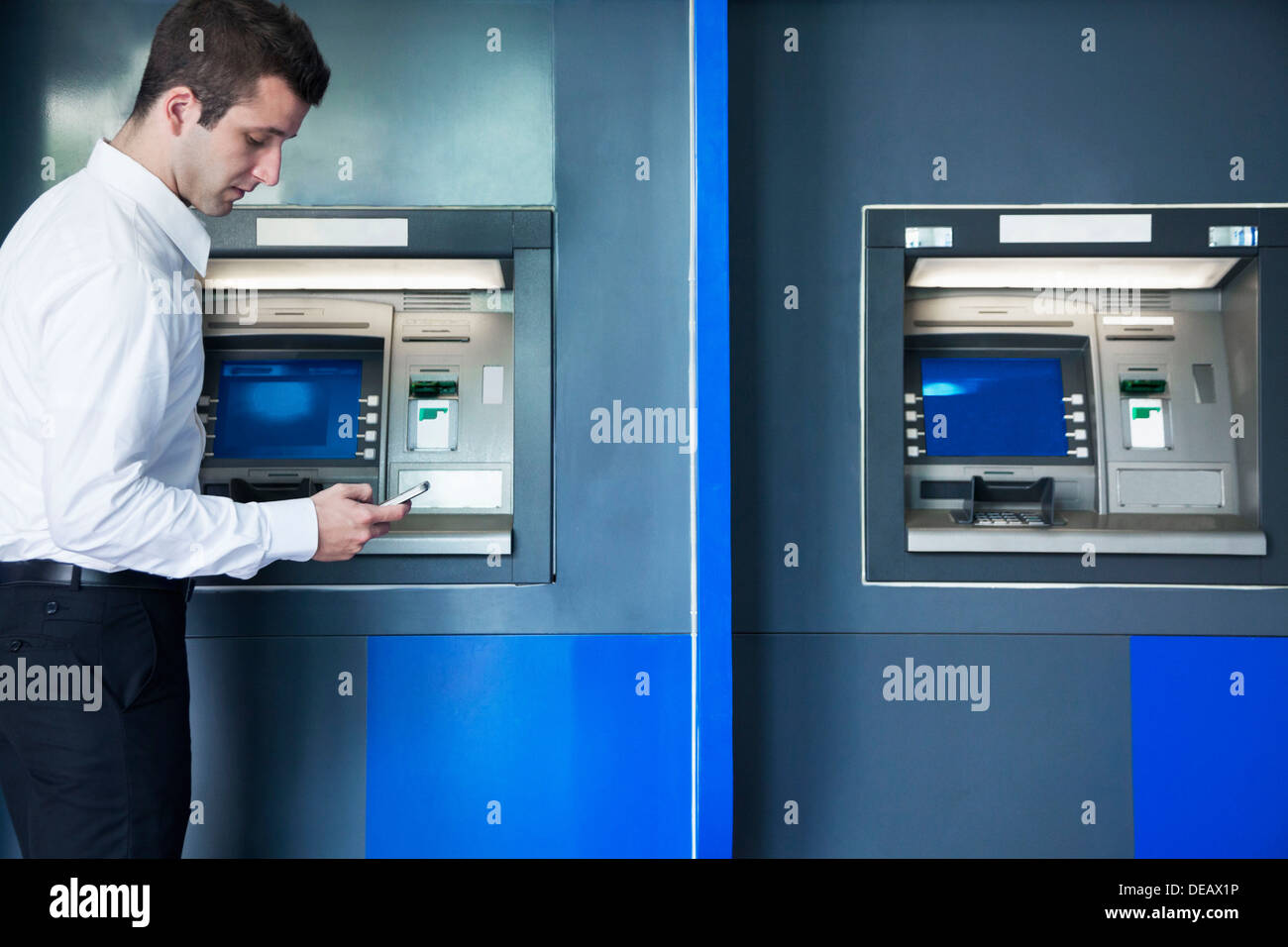 Young businessman taking out money from the ATM and looking down at his phone Stock Photo