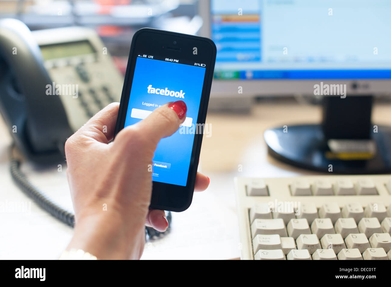 Woman's hand holding a smart phone with facebook on the screen in an office with a desk, phone, computer screen and keyboard Stock Photo