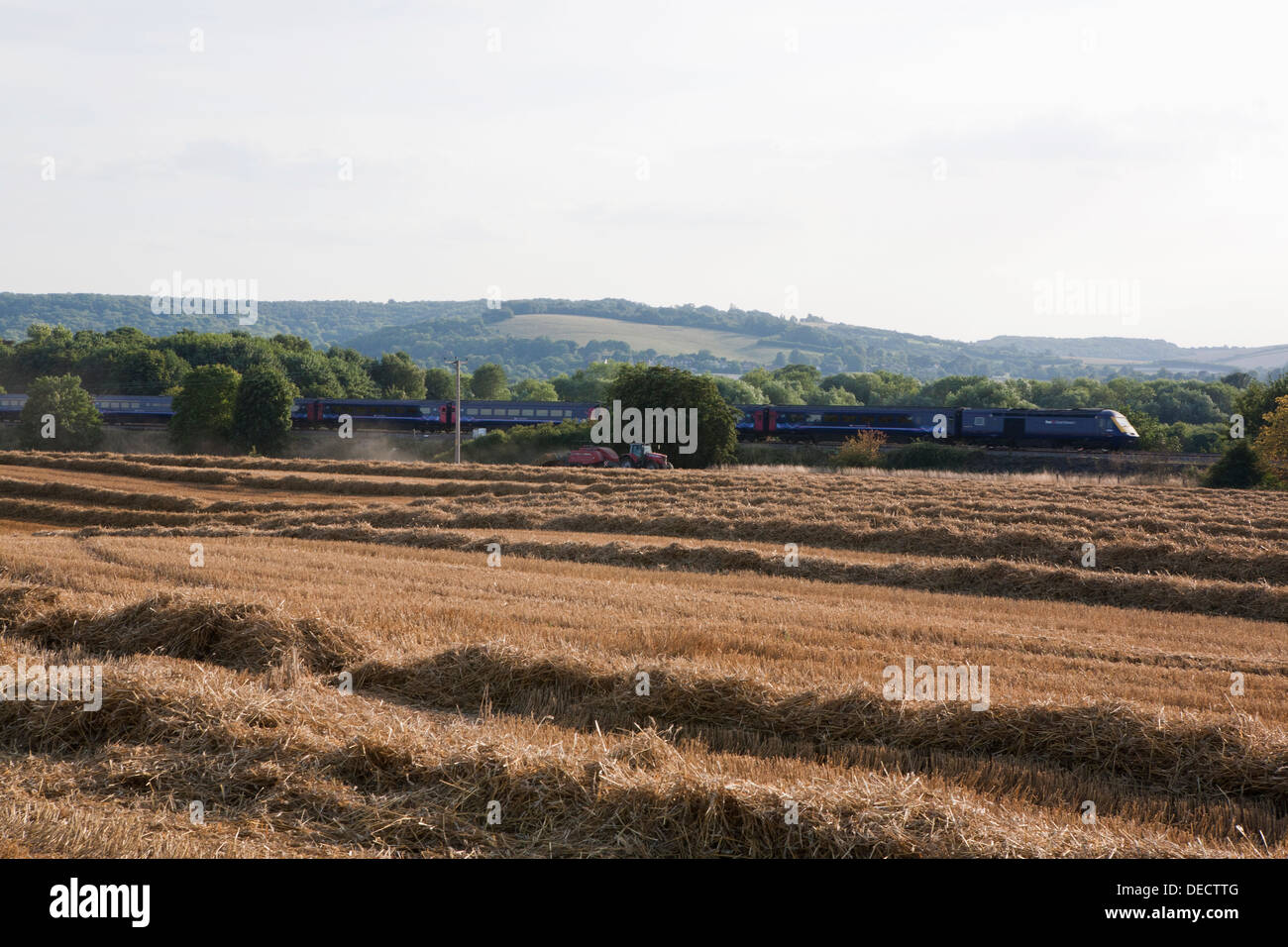 A red Massey Ferguson tractor and straw baler operating in a harvested field. A train passes by at high speed. Stock Photo