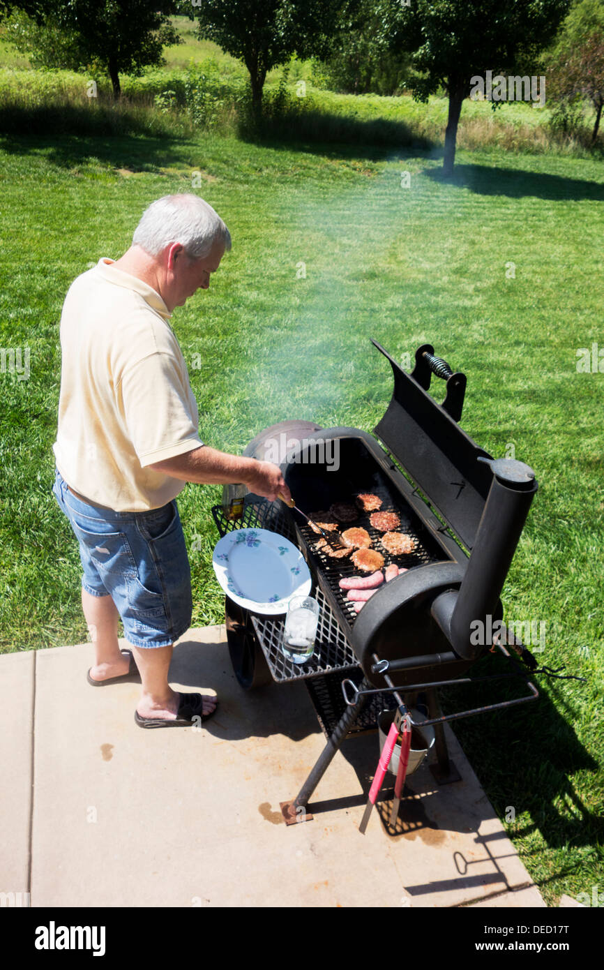 A man in his late 40s grills hamburgers and brats on a smoker/grill in his back yard. Wichita, Kansas, USA. Stock Photo