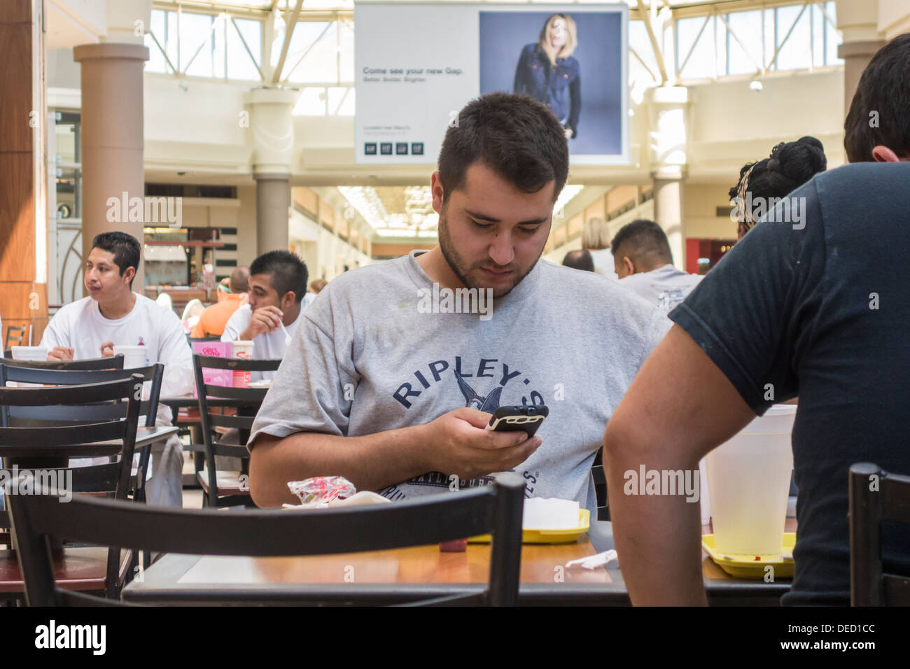A young man in his mid 20s is engrossed in his cell phone while eating in a food court in a shopping mall. Oklahoma City, Oklahoma, USA. Stock Photo