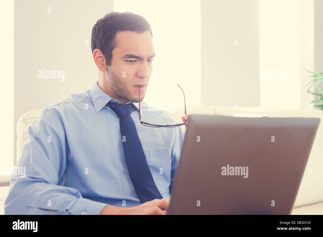 Frowning man biting his eyeglasses and using a laptop Stock Photo