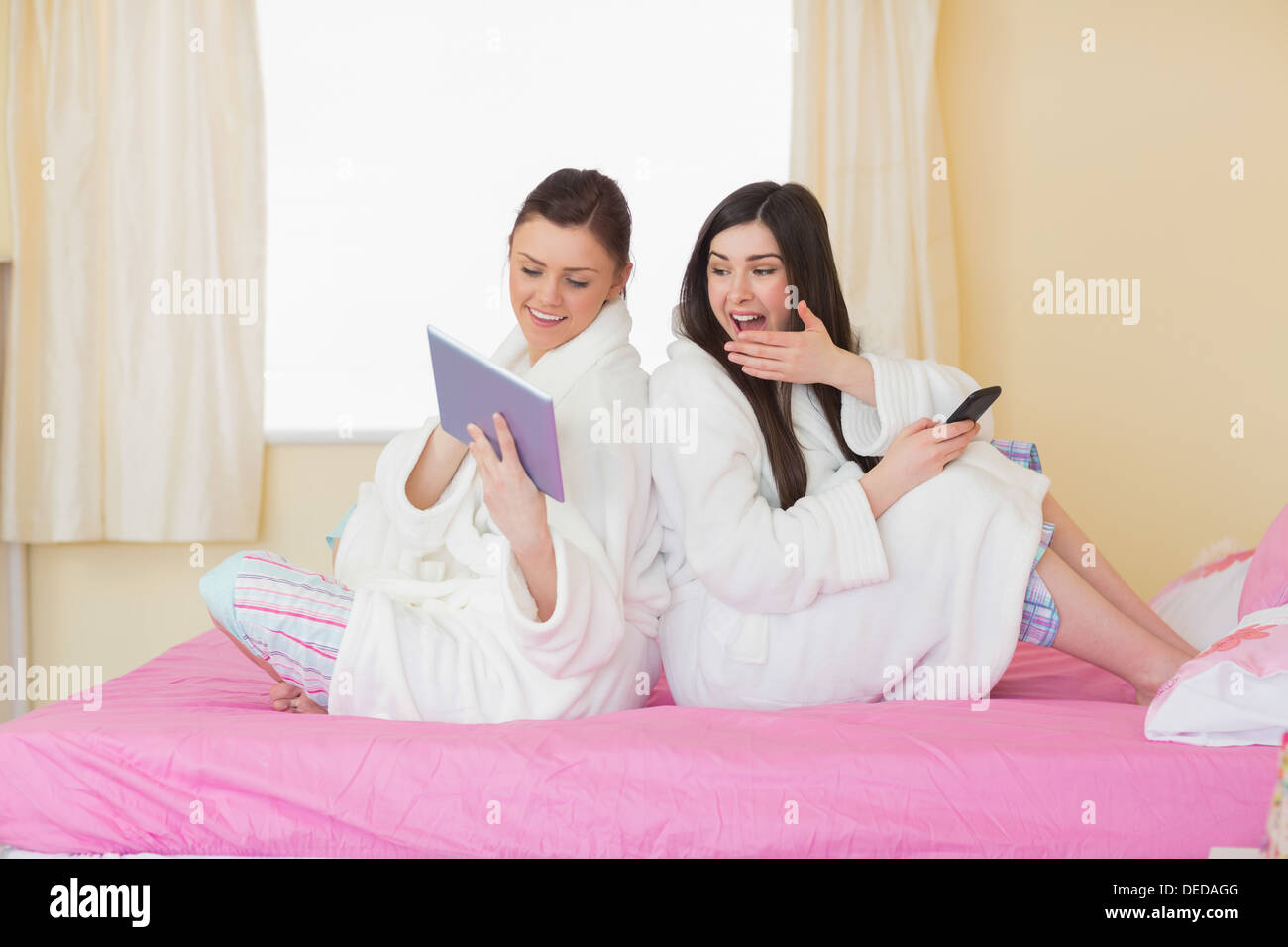 Two friends wearing bathrobes sitting back-to-back looking at tablet Stock Photo