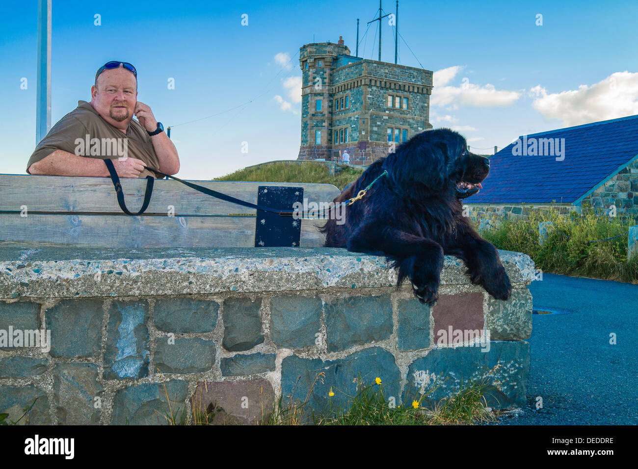 A middle-aged man sits with his black Newfoundland dog by a stone wall on top of 'Signal Hill' in St. John's. Newfoundland. Stock Photo