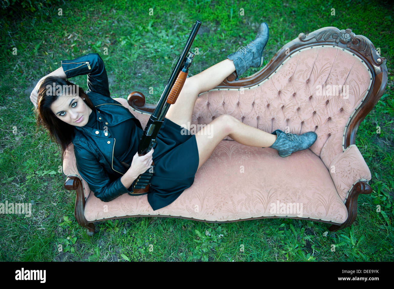 Sexy Caucasian female model posing in a seductive, yet flattering manner on a vintage couch with a shotgun, boots & jacket. Stock Photo