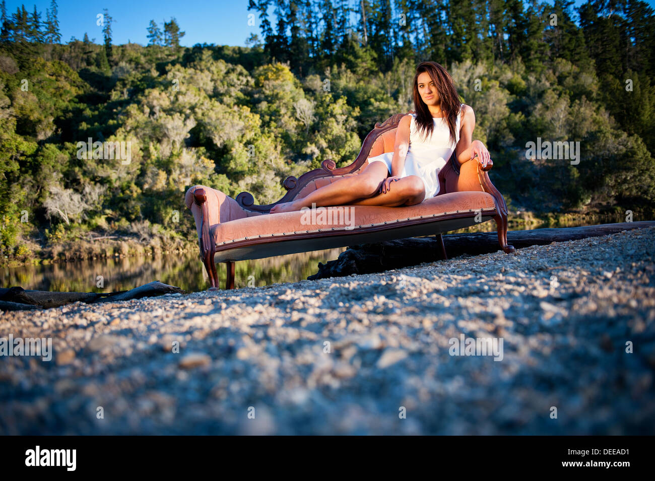 Attractive young Caucasian female model posing on a beautiful vintage couch outside a remote natural environment during sunset. Stock Photo