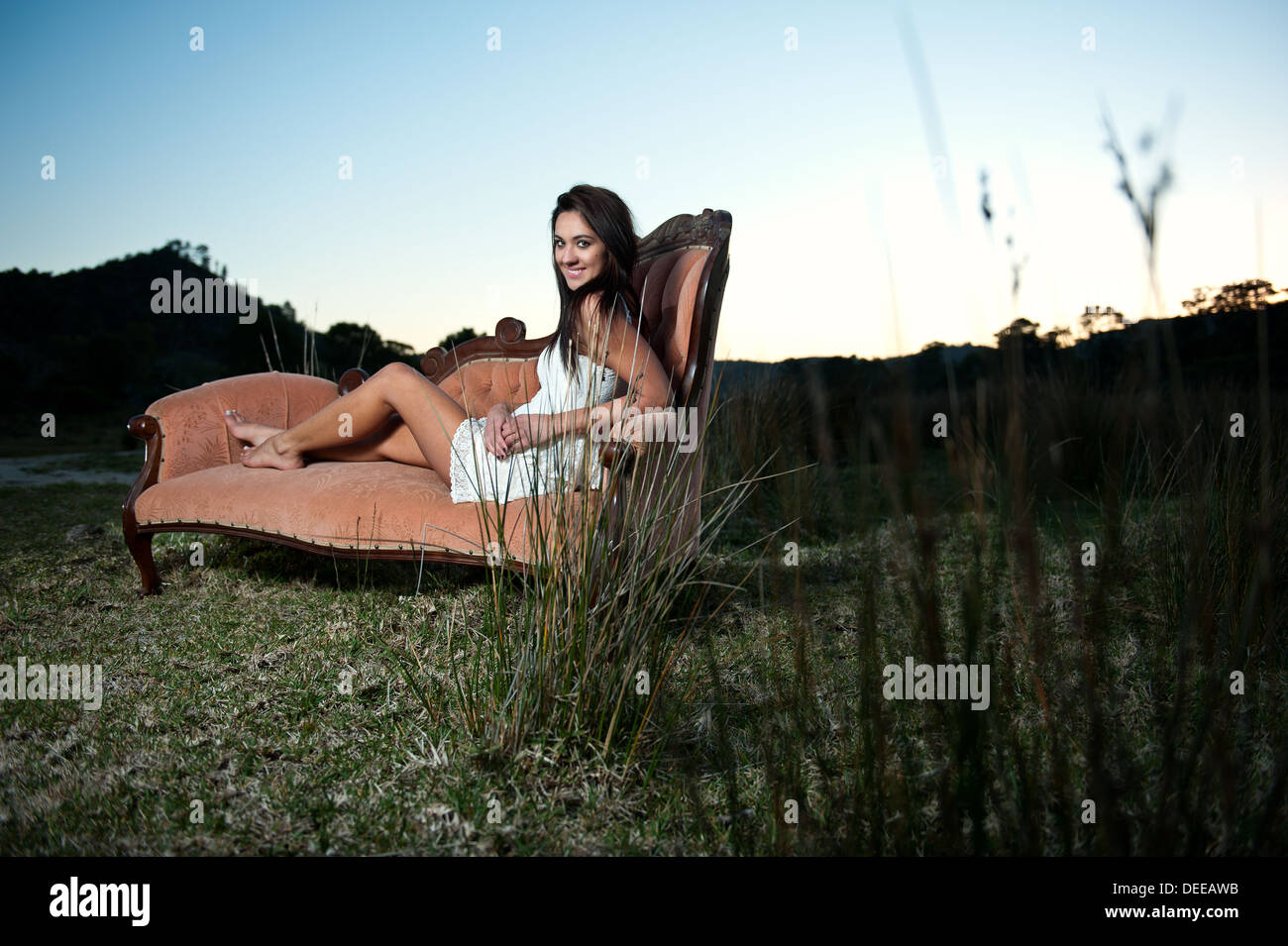 Attractive young Caucasian female model posing on a beautiful vintage couch outside a remote natural environment during sunset. Stock Photo