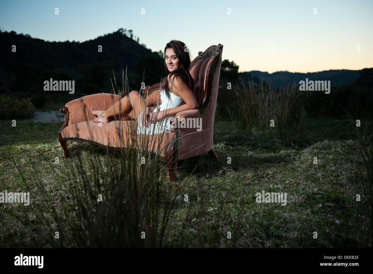 Attractive young Caucasian female model posing on a beautiful vintage couch outside a remote natural environment during sunset. Stock Photo