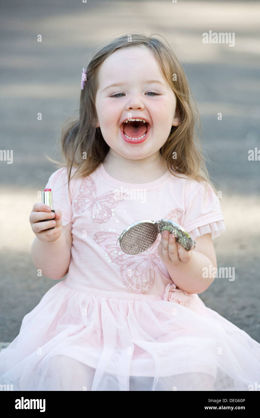 A pretty 2 year old girl wearing a pink dress and playing with lipstick. Stock Photo