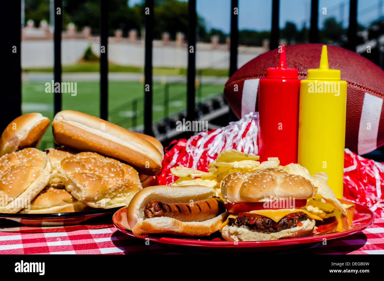 Football party with cheeseburger, hot dog, potato chips, pom poms, buns, and football. Football field in background. Stock Photo