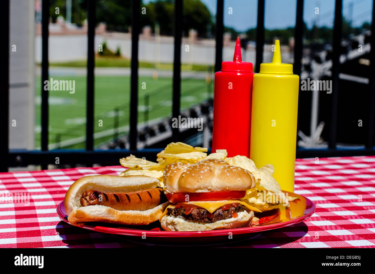 Tailgate party with cheeseburger, hot dog, potato chips and mustard and ketchup bottles. Football field in background. Stock Photo