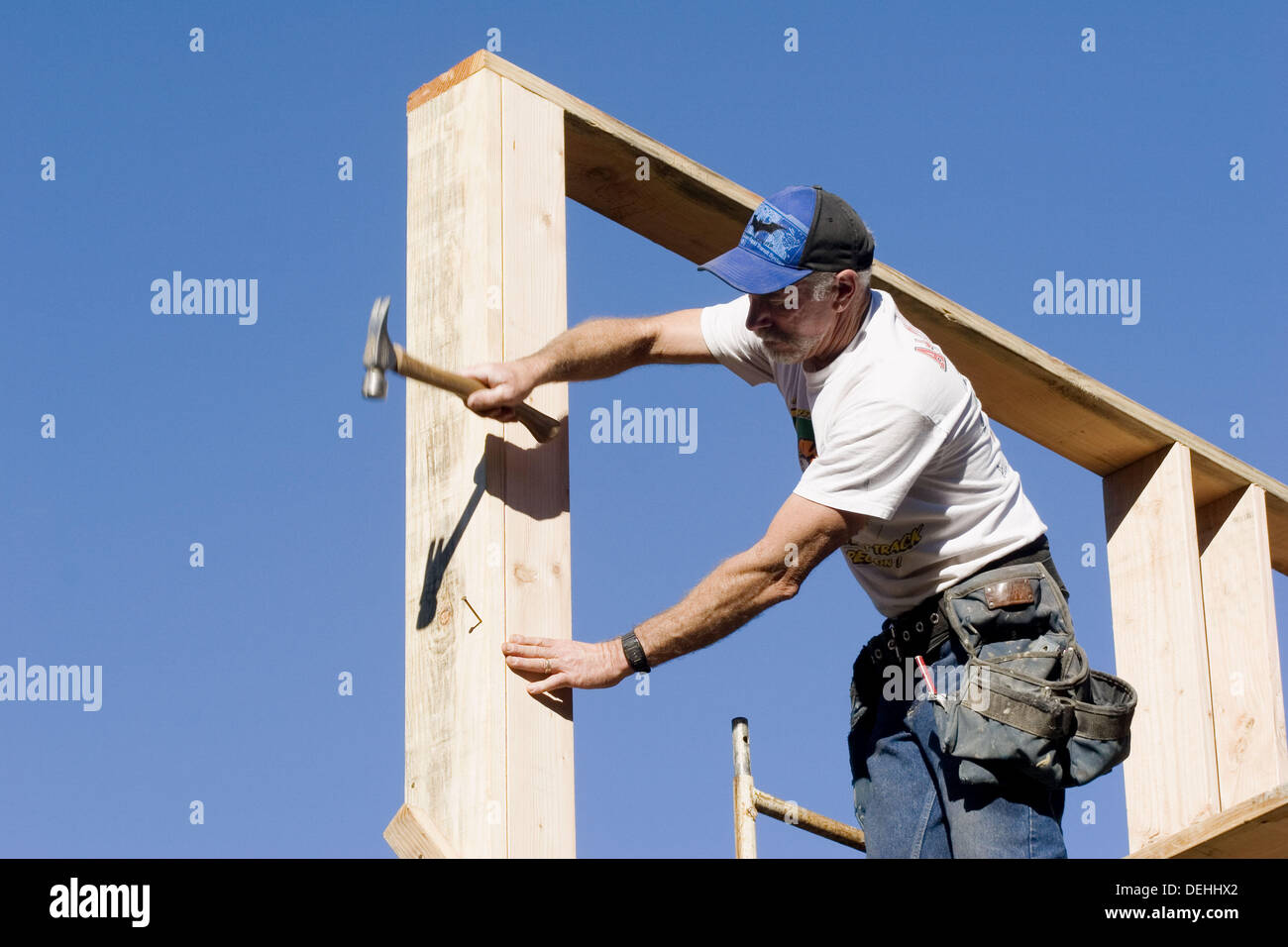 Carpenter reaching to hammer a nail into the corner stud of a twostory room while standing on
