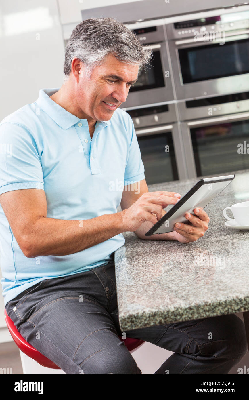 Middle aged man using tablet computer at home in the kitchen drinking coffee or tea Stock Photo