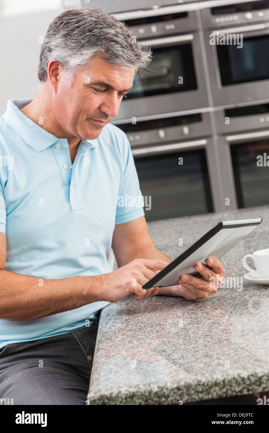 Middle aged man using tablet computer at home in the kitchen drinking coffee or tea Stock Photo
