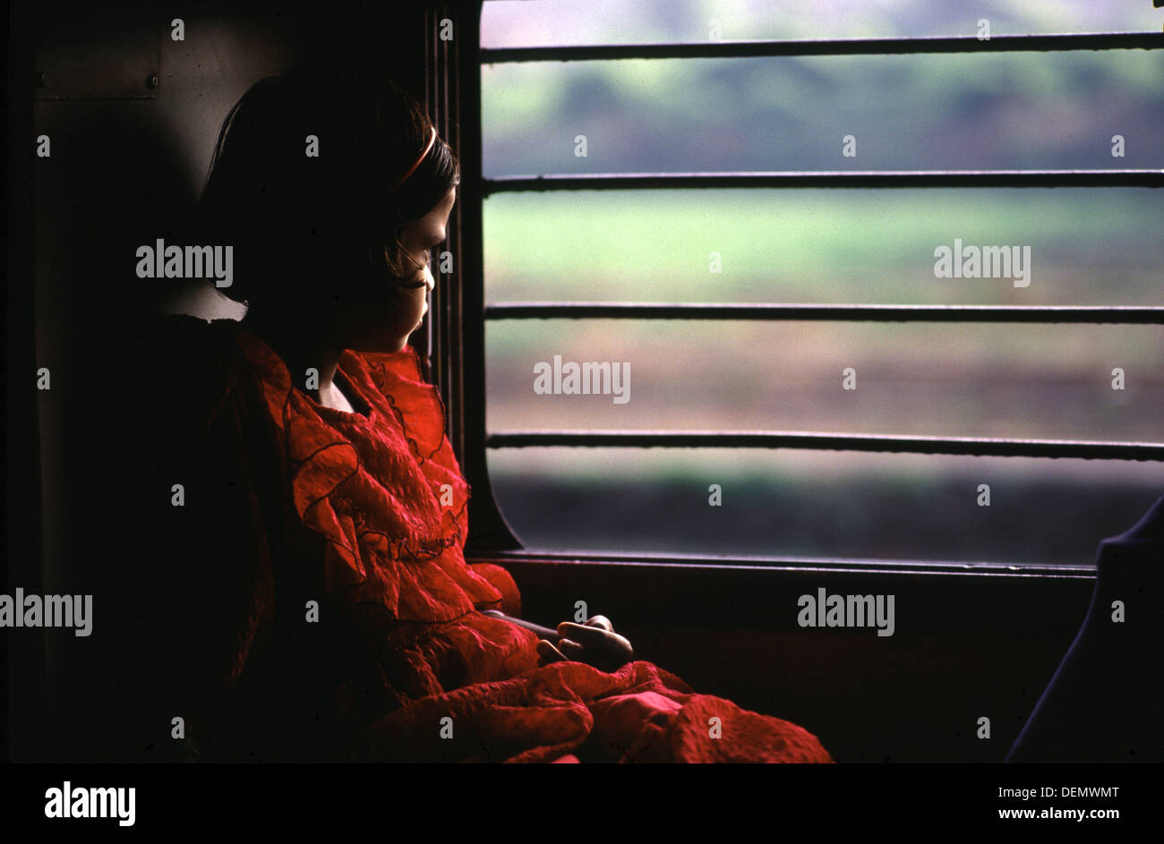 A young Indian girl wearing a red dress watching countryside roll by from a train in India Stock Photo