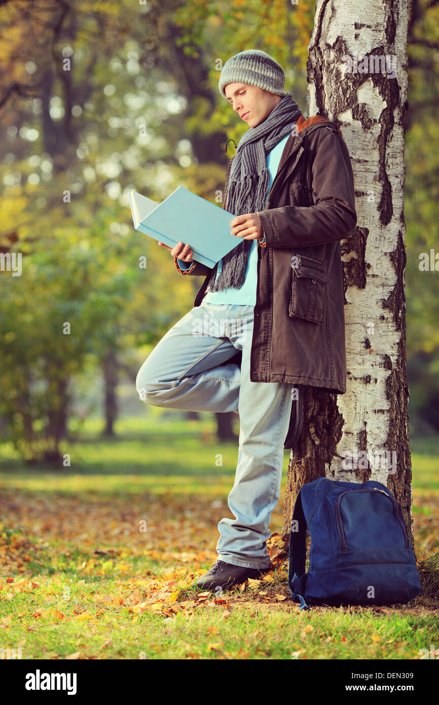 Full length portrait of a male student leaning on a tree and reading a book in a park Stock Photo