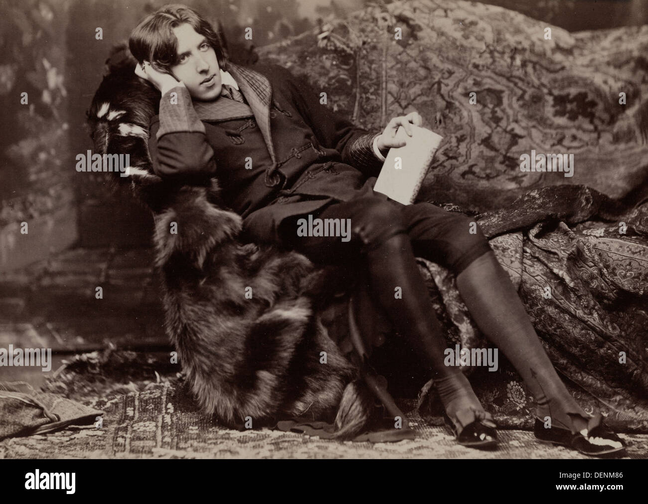 Oscar Wilde, full-length portrait, facing right, sitting in chair, right hand on cheek, left hand holding book. 1882 Stock Photo