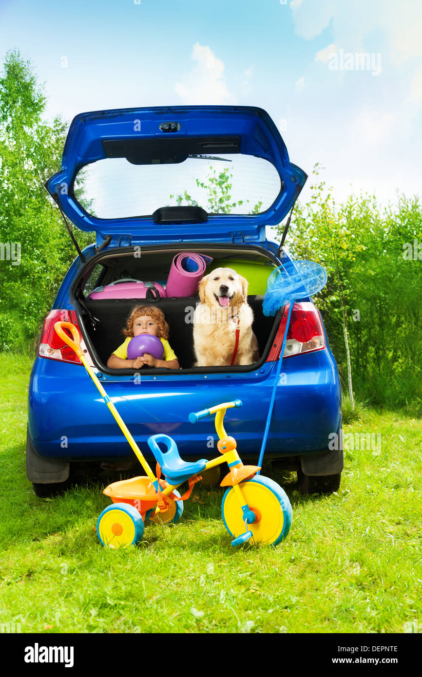 Little three years old boy sitting in the car trunk with a dog waiting for parents to put bags and tricycle for the trip in the car Stock Photo