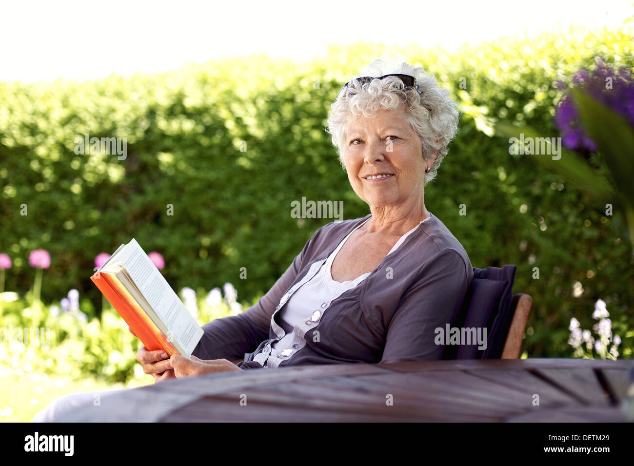 Happy senior woman with book in hand sitting in her backyard looking at camera smiling - Elder woman reading novel in garden Stock Photo