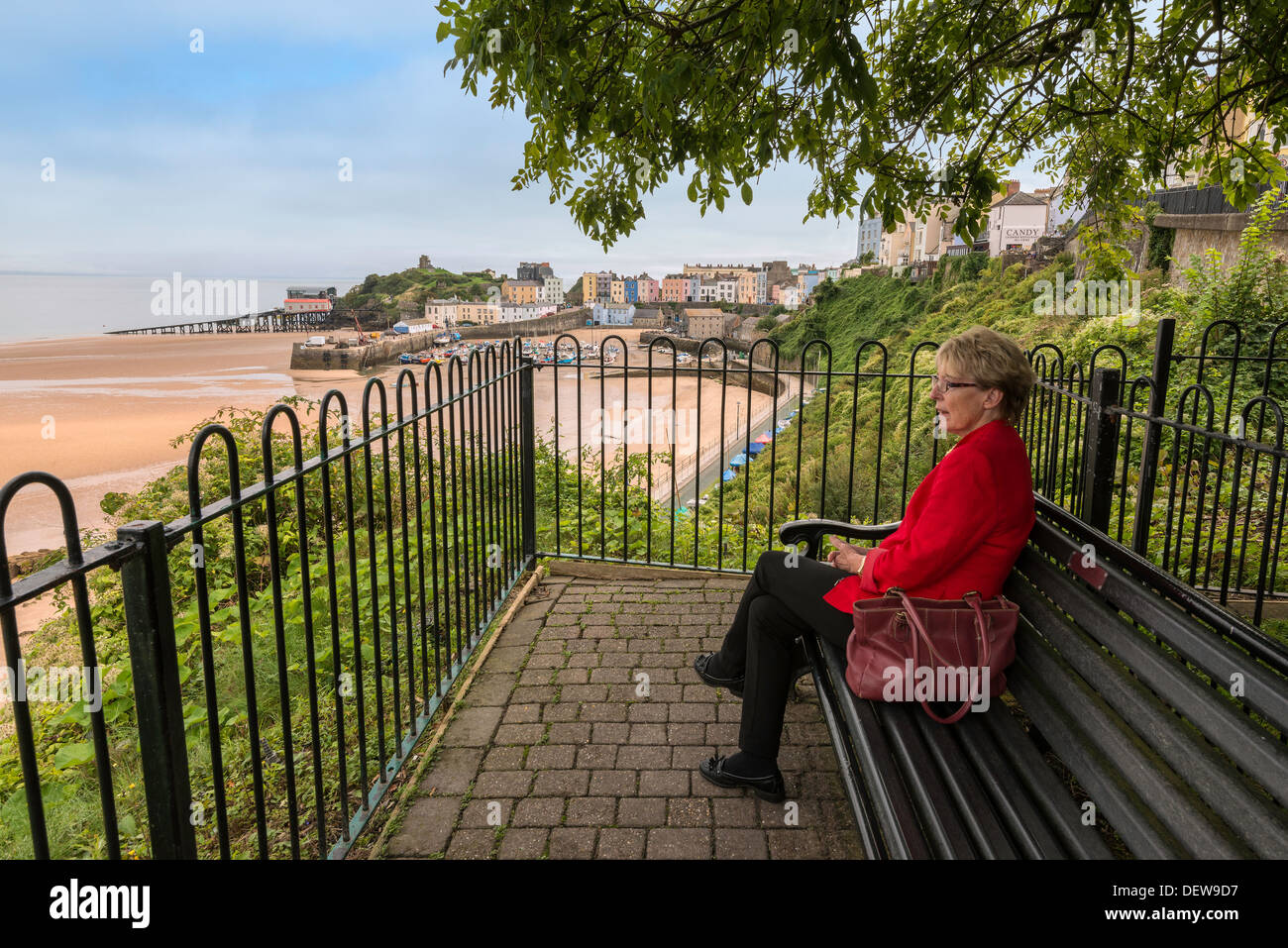Older woman sitting on bench overlooking North Beach, Tenby Pembs.Low tide with moored boats and terraces of Regency type houses Stock Photo