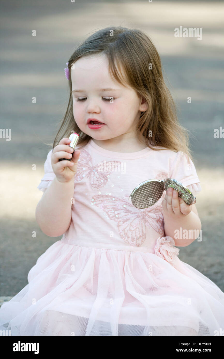 A pretty 2 year old girl wearing a pink dress and playing with lipstick. Stock Photo