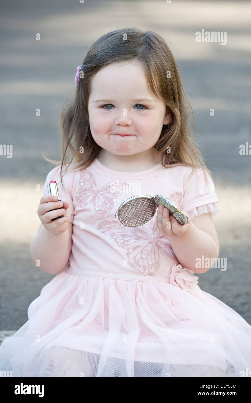 A pretty 2 year old girl wearing a pink dress and playing with lipstick. Stock Photo