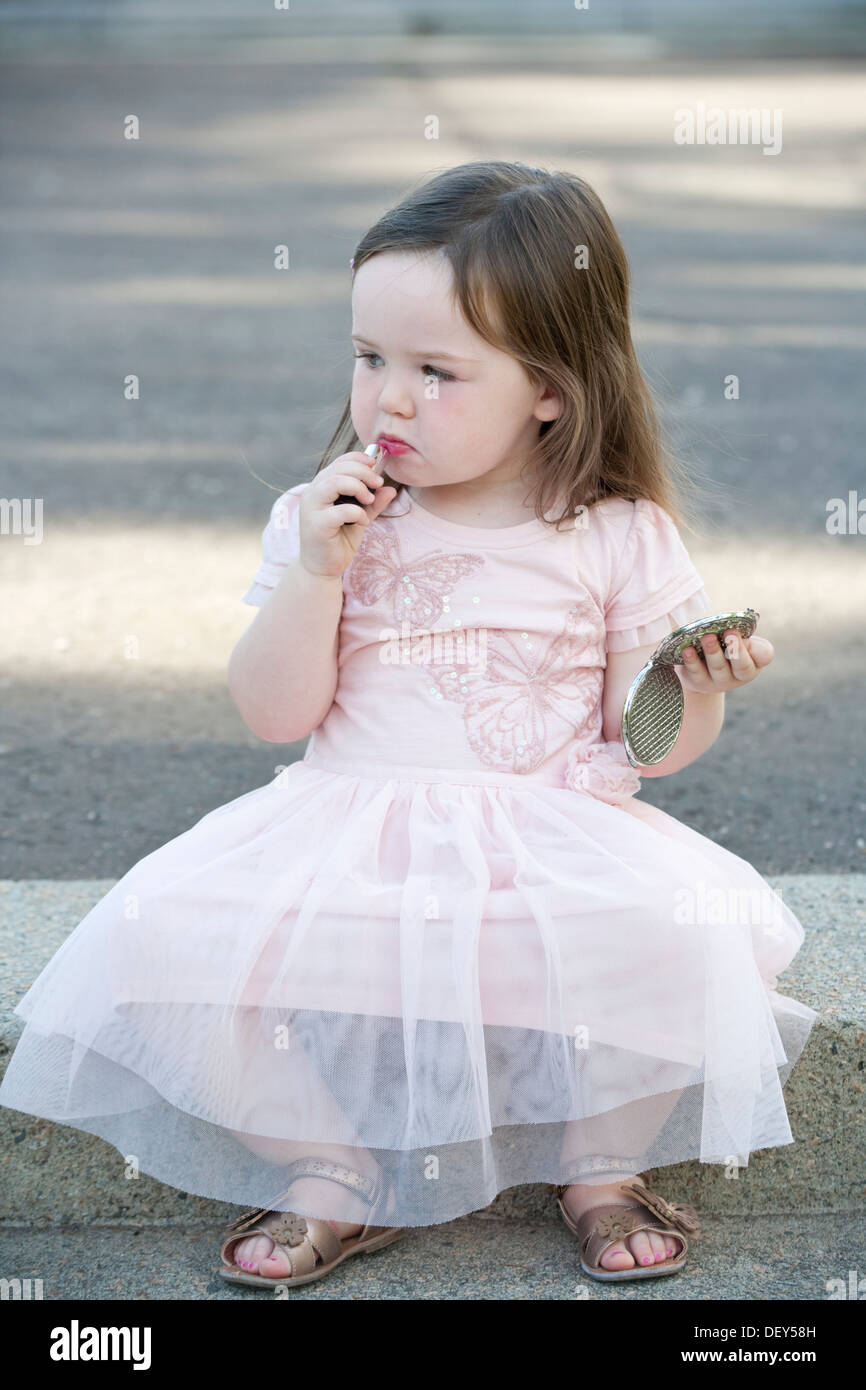 A pretty 2 year old girl wearing a pink dress and playing with lipstick. Stock Photo