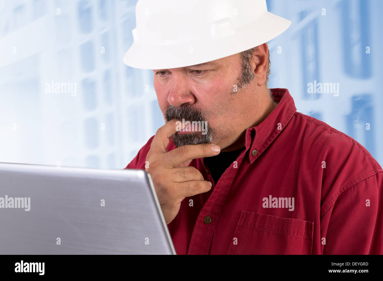 Hardhat worker perplexed, working on the tough issues with his laptop, he has a serious thoughtful look that he values his job Stock Photo
