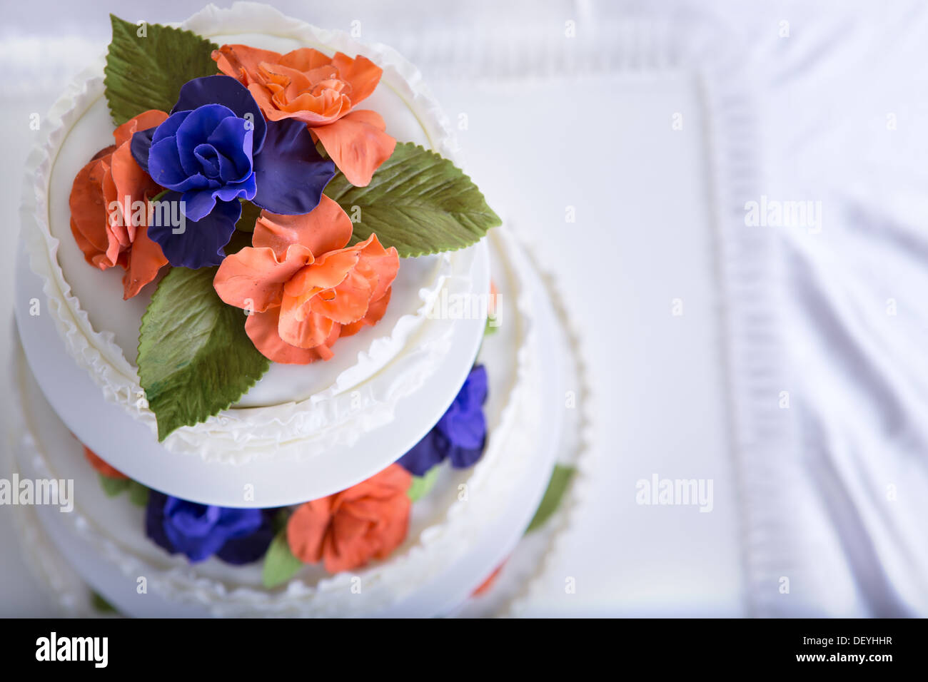 Three tier fondant ruffled white wedding cake decorated with orange and purple gum paste roses and green leafs, shot from above Stock Photo