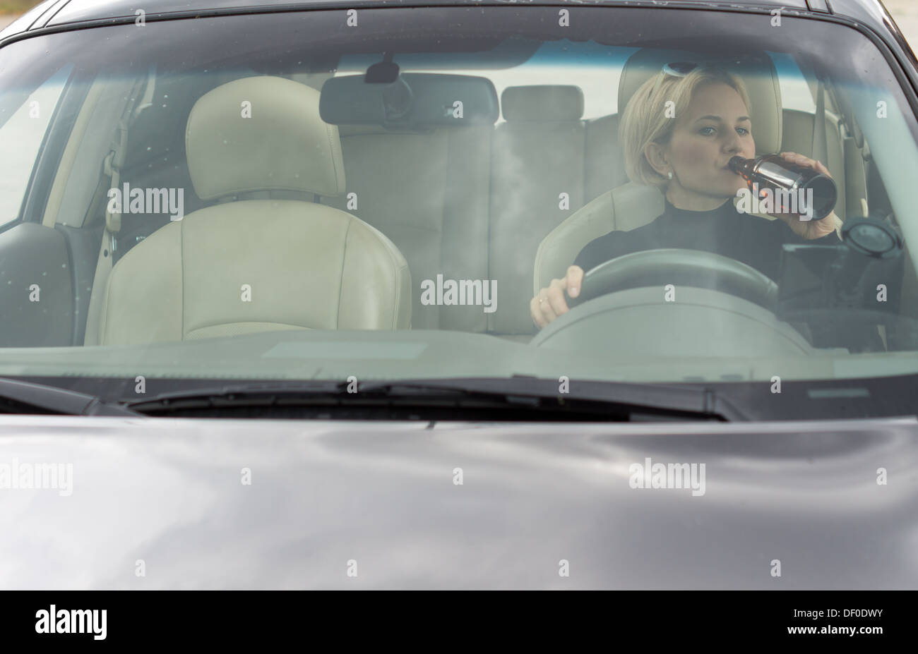 Female alcoholic sitting in her car drinking and driving as she gulps alcohol directly from the bottle, view through the front windscreen. Stock Photo