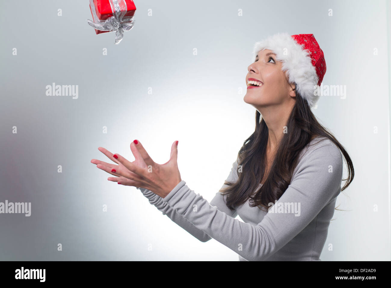 Laughing beautiful woman wearing a red Santa hat catching a surprise Christmas gift suspended midair above her outstretched hand Stock Photo