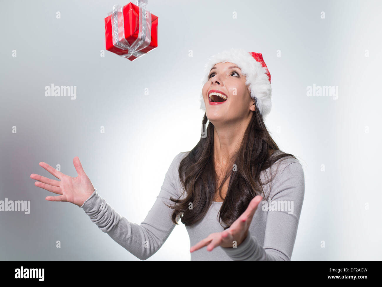 Laughing beautiful woman wearing a red Santa hat catching a surprise Christmas gift suspended midair above her outstretched hand Stock Photo
