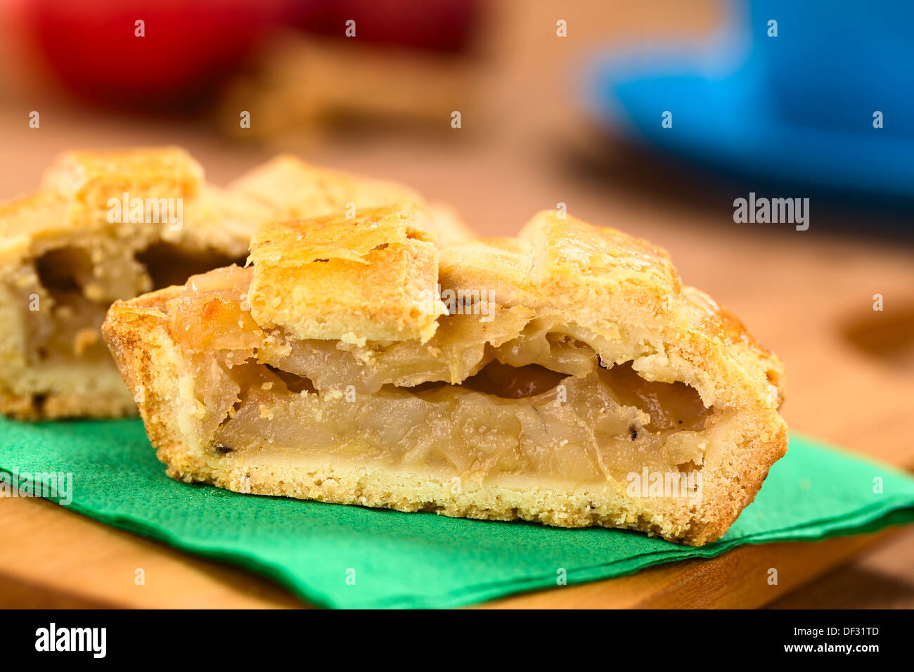 Half apple pie with lattice crust on green napkin on wooden board, with a blue cup, apples and cinnamon sticks in the back Stock Photo