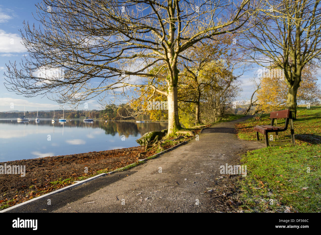 A serene scene of a bench in a lake-side park near Ambleside on a sunny morning with mirror-like water and yachts. Stock Photo