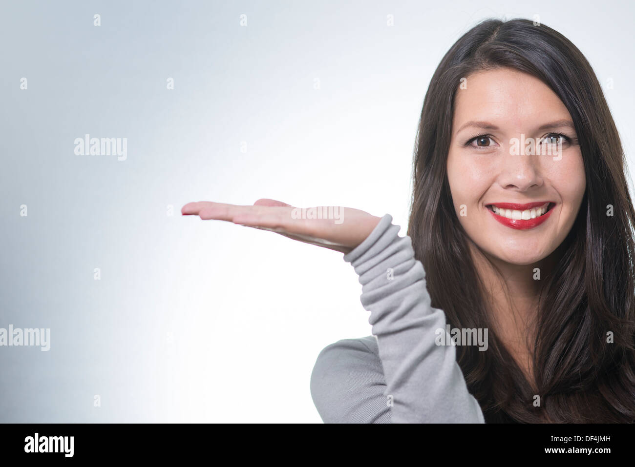 Beautiful young woman with a lovely friendly smile standing with an outstretched empty palm for your product placement pointing Stock Photo