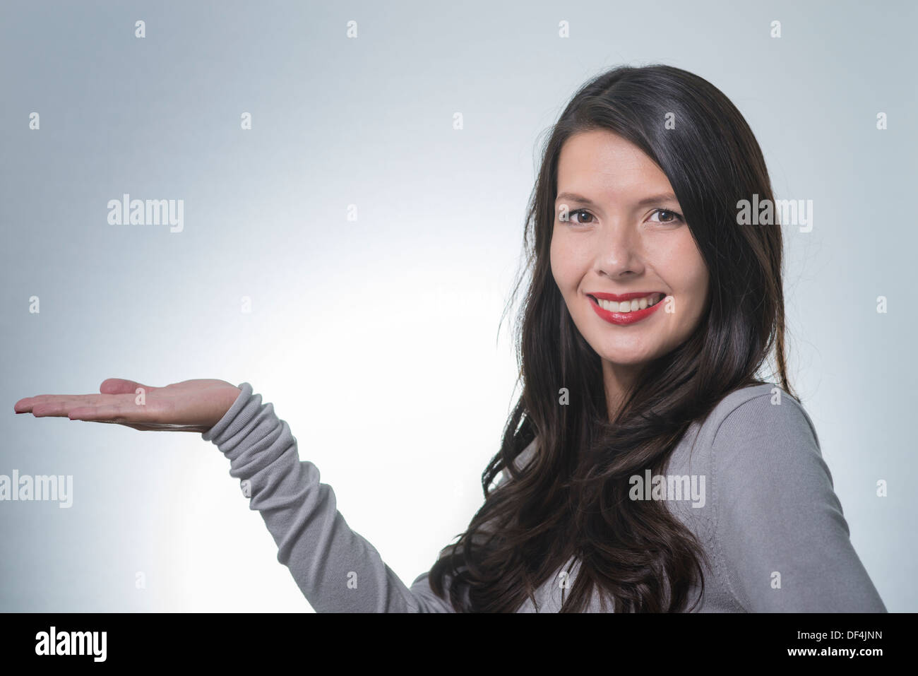 Beautiful young woman with a lovely friendly smile standing with an outstretched empty palm for your product placement pointing Stock Photo