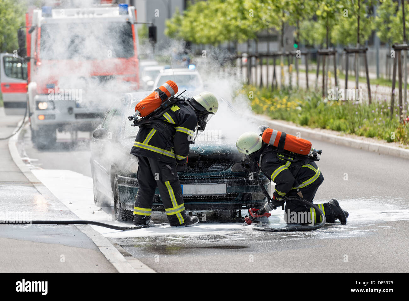 Burning motor vehicle been put out by firemen in protective clothing Stock Photo