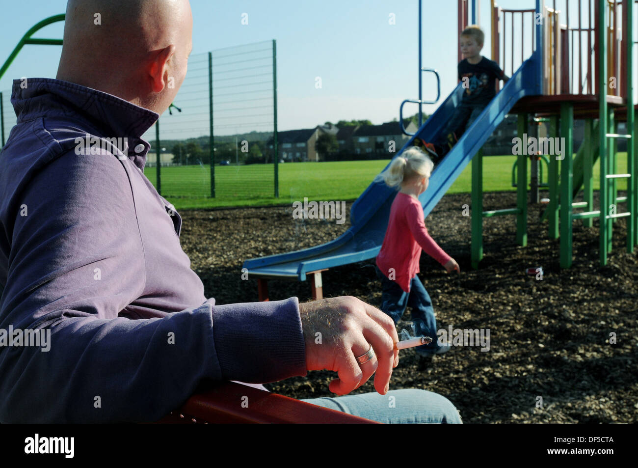 man smoking in a children's playground with a girl and boy playing on a slide Stock Photo