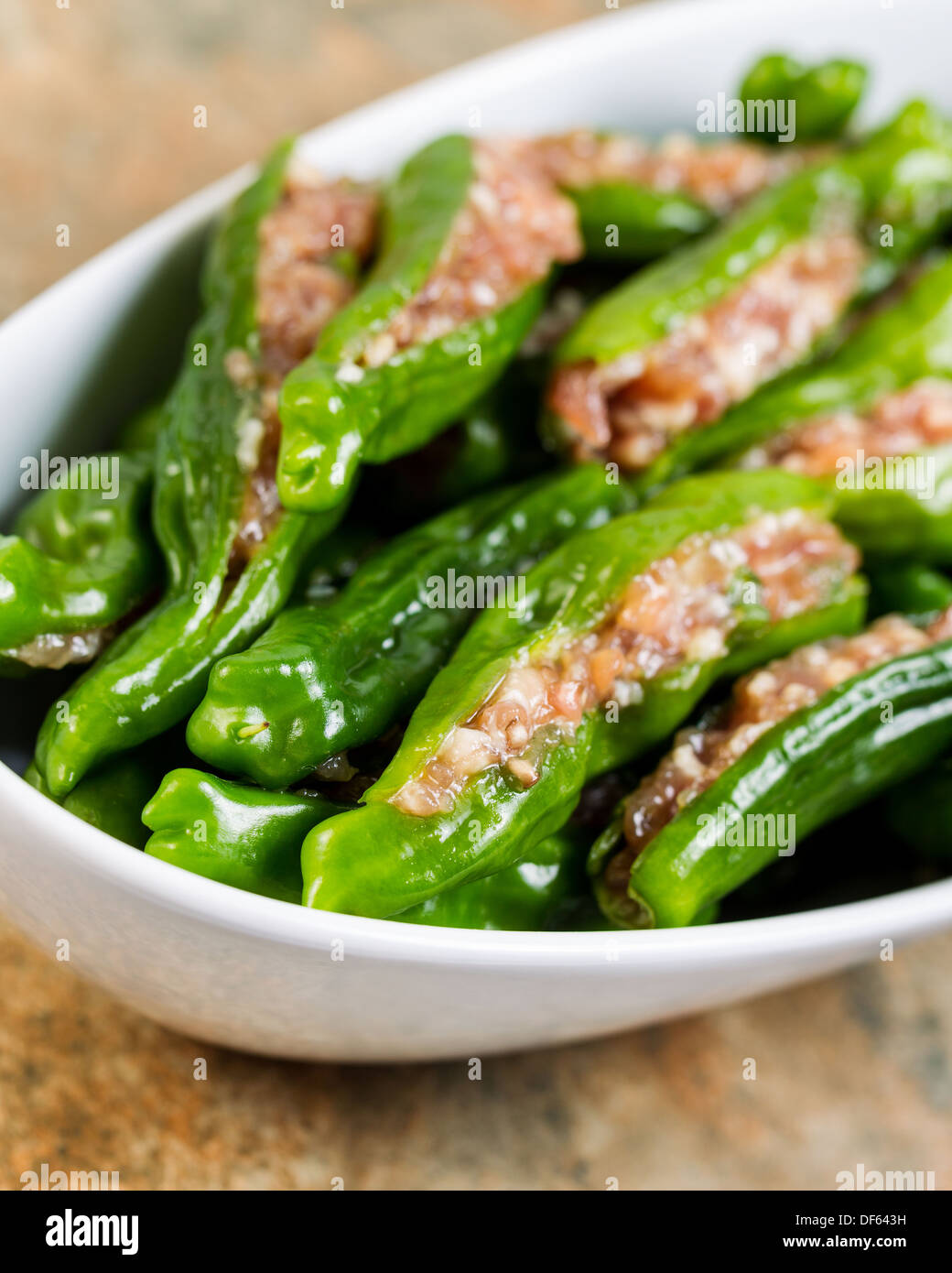Closeup vertical photo of uncooked fresh, stuffed green sweet peppers, in white bowl, on stone counter top Stock Photo