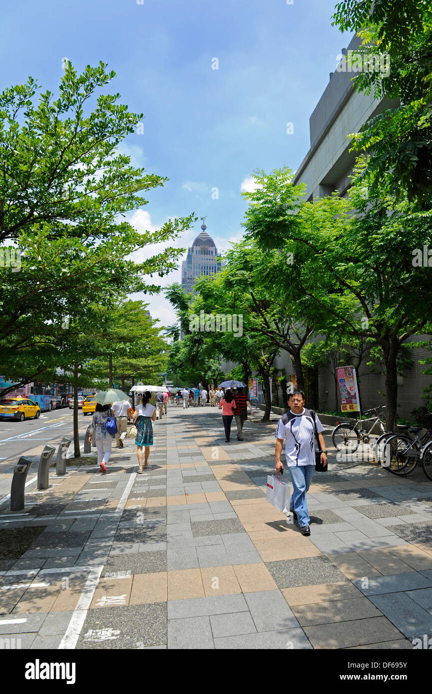 Male and female pedestrians walking along a wide tree lined pavement on a sunny in the capital city of Taiwan, Taipei. Stock Photo