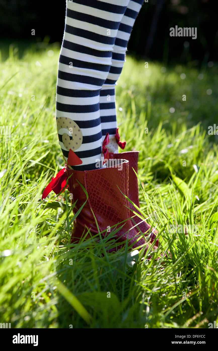 Side shot of a child's legs in stripy tights and red boots standing in the long overgrown green grass of a meadow. Stock Photo