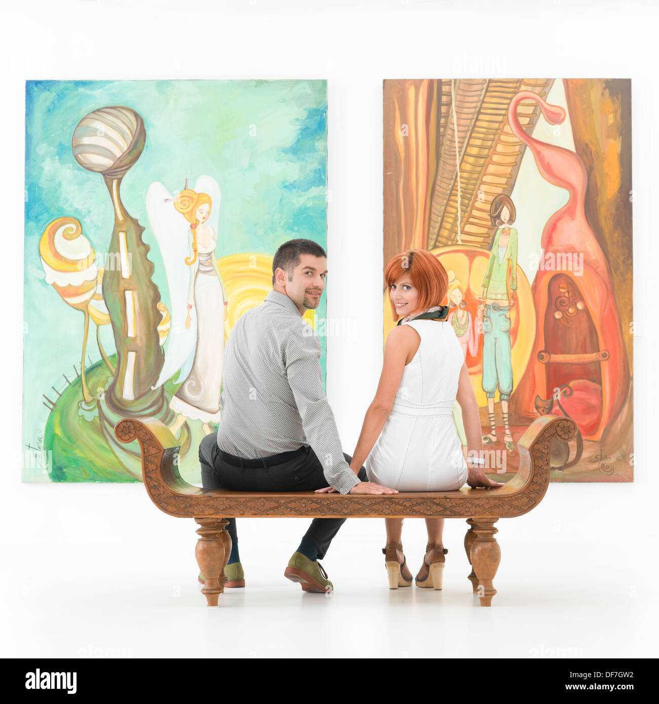 young couple sitting on a wooden bench in front of large colorful paintings holding hands and smiling Stock Photo