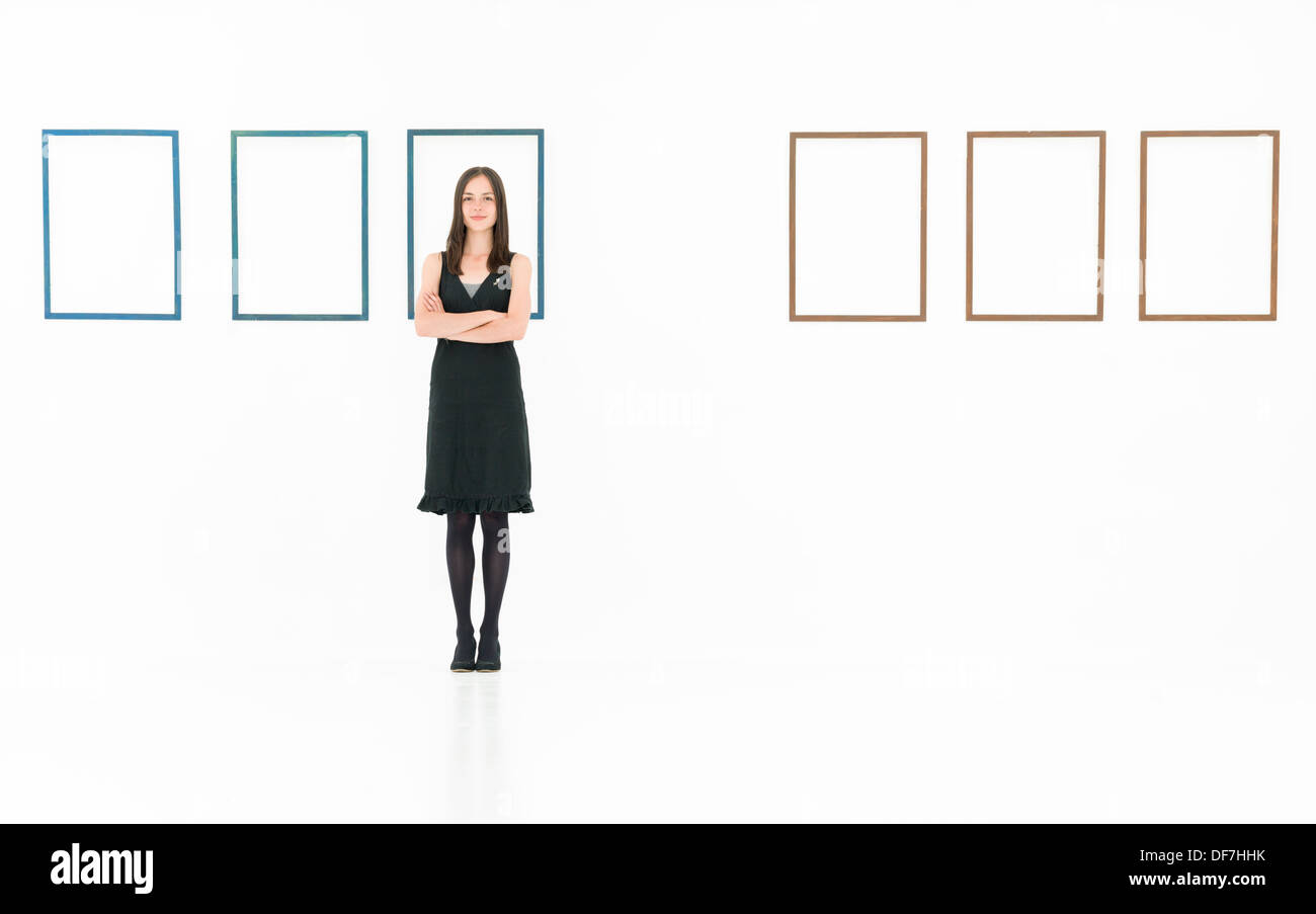 front view of woman standing in a white room with arms crossed with empty frames on a wall behind her Stock Photo