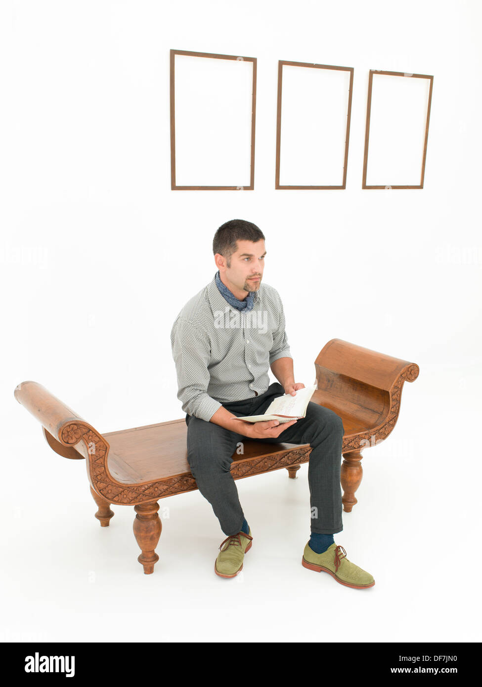 young caucasian man sitting on a brown wooden bench holding a book, in front of empty frames displayed on white wall Stock Photo