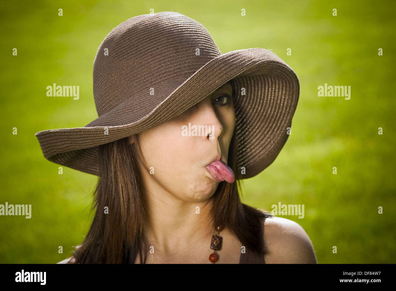 Young Spanish/Asian woman, wearing a hat, sticking out her tongue while posing at a park Stock
