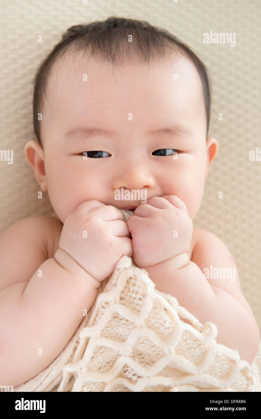 Adorable six months old teething Asian baby girl lying on bed biting blanket. Stock Photo