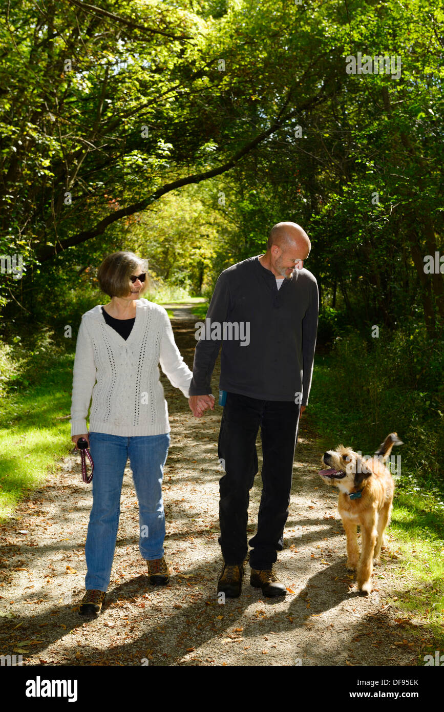 Smiling couple holding hands walking up a woodland path with an attentive pet dog off leash in a Toronto park Stock Photo